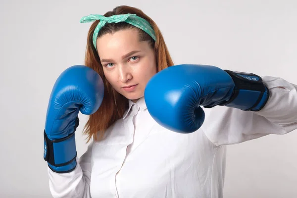 Girl power concept. Confident young woman isolated on a gray wall background. Female and independent power. The girl raises her hand in a boxing glove to strike. Studio shot.