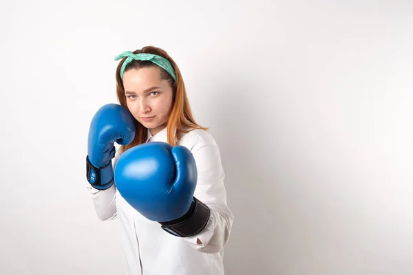 Girl power concept. Confident young woman isolated on a gray wall background. Female and independent power. The girl raises her hand in a boxing glove to strike. Studio shot.