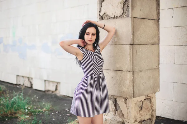 young skinny tall woman posing in short dress with stripes. nice, charming and sexy brunette shows fashion pose on the background of the abandoned buildings