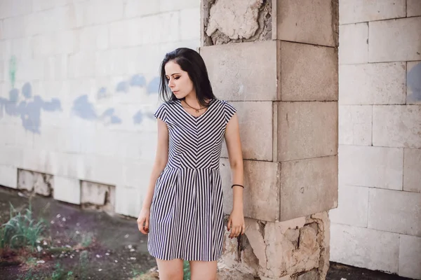 young skinny tall woman posing in short dress with stripes. nice, charming and sexy brunette shows fashion pose on the background of the abandoned buildings