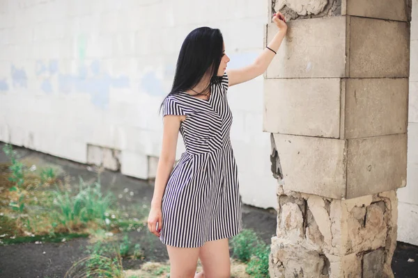 Junge schlanke, große Frau, die in einem kurzen Kleid mit Streifen posiert. schöne, charmante und sexy Brünette zeigt Mode-Pose auf dem Hintergrund der verlassenen Gebäude — Stockfoto
