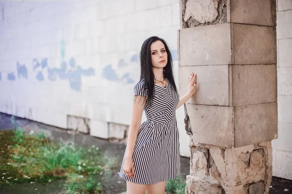 young skinny tall woman posing in short dress with stripes. nice, charming and sexy brunette shows fashion pose on the background of the abandoned buildings