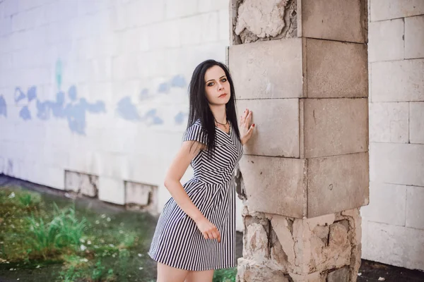 Ung smal lång kvinna poserar kort klänning med ränder. fin, charmig och sexig brunett visar mode pose på bakgrunden av de övergivna byggnaderna — Stockfoto