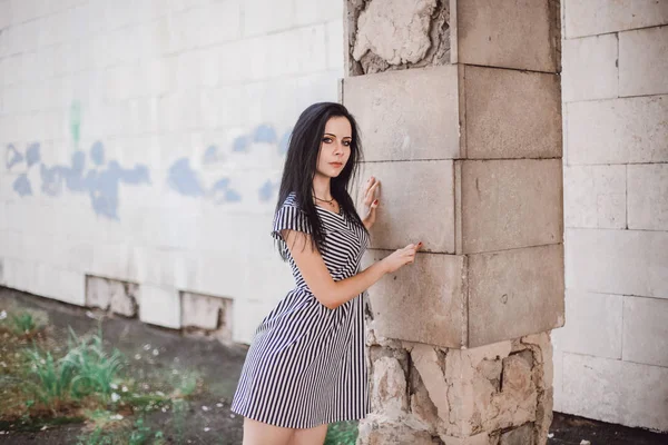 young skinny tall woman posing in short dress with stripes. nice, charming and sexy brunette shows fashion pose on the background of the abandoned buildings