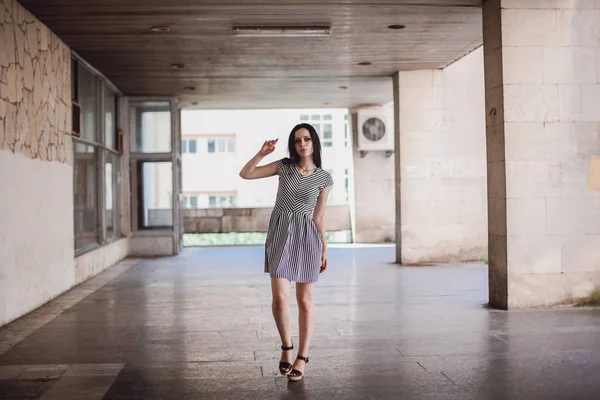 young skinny tall woman posing in short dress with stripes. charming and sexy brunette shows fashion pose on the background of the abandoned buildings.
