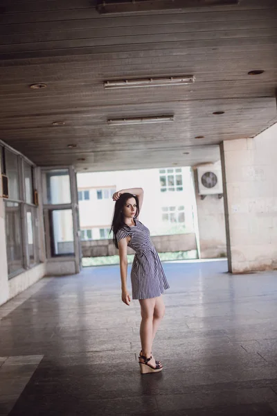 Junge schlanke, große Frau, die in einem kurzen Kleid mit Streifen posiert. Charmant und sexy Brünette zeigt Mode Pose auf dem Hintergrund der verlassenen Gebäude. — Stockfoto