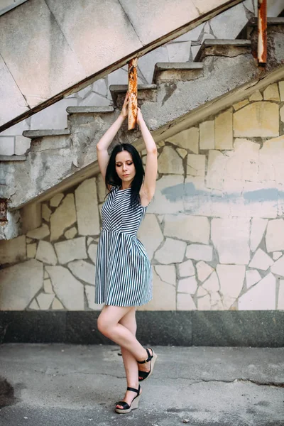 young skinny charming lady posing in short dress black and white with stripes. nice and sexy brunette demonstrating fashion pose on the background of the abandoned buildings