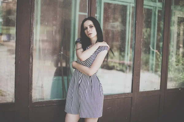 young skinny charming lady posing in short dress black and white with stripes. nice and sexy brunette demonstrating fashion pose on the background of the abandoned buildings