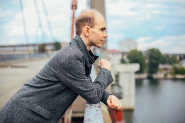 uzağa arıyorsunuz köprüde duran ve poz moda vogue, bulutlu ve soğuk rüzgarlı hava, arka plan şehir, genç adam portresi. uzun gri ceket şık