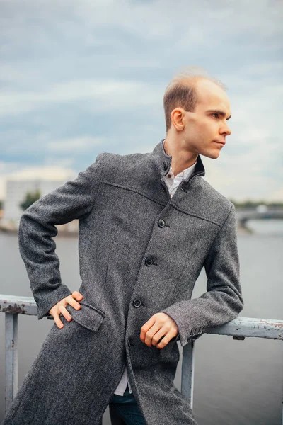 portrait of young man standing on the bridge and posing fashion vogue, cloudy and cold windy weather, background city, looking away. in a long gray coat stylish