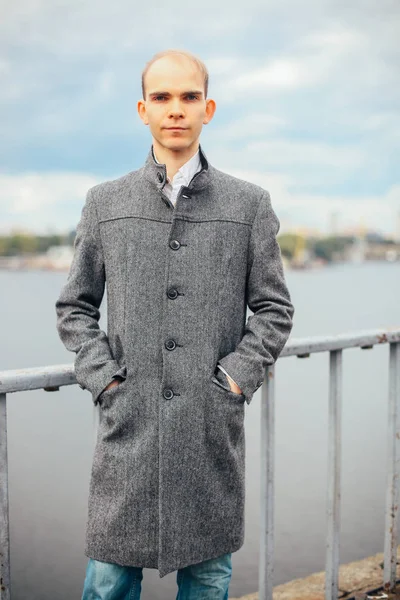 portrait of young man standing on the bridge and posing fashion vogue, cloudy and cold windy weather, background city, looking away. in a long gray coat stylish