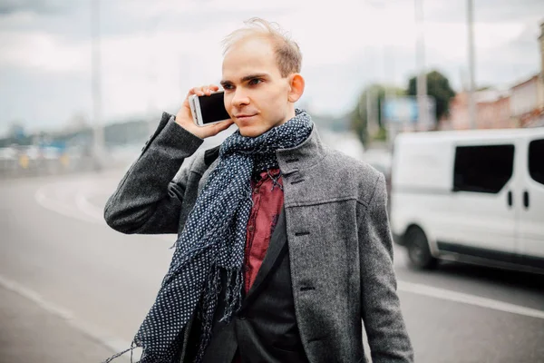 portrait of young man standing near the road with heavy traffic and posing, cloudy and cold windy weather closeup, talking on the phone with business partners, the wind blows long stylish gray coat