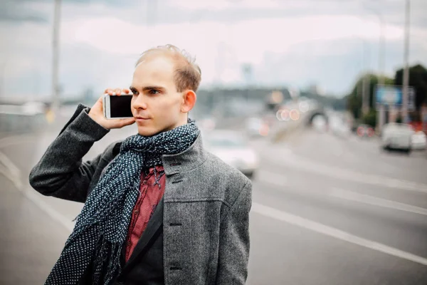 portrait of young man standing near the road with heavy traffic and posing, cloudy and cold windy weather closeup, talking on the phone with business partners, the wind blows long stylish gray coat