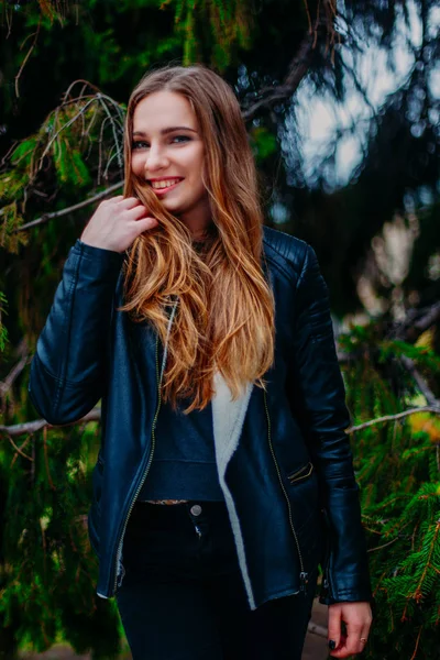 Potret seorang wanita muda berambut pirang yang cantik hipster dengan jaket bulu hitam berpose di cabang-cabang pohon cemara. tersenyum kebahagiaan wanita beruntung, musim gugur dingin atau musim semi — Stok Foto