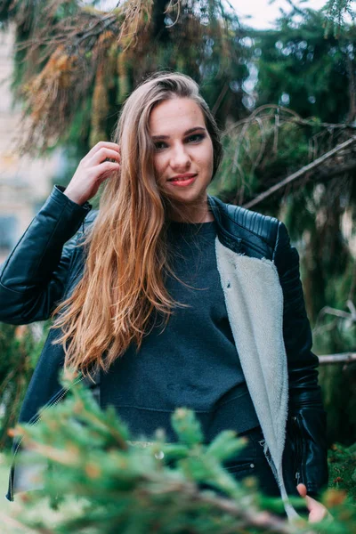 Porträt einer schönen blonden jungen kaukasischen Hipsterin in einer schwarzen Pelzjacke, die modisch in Tannenzweigen posiert. lächelnde glückliche Frau, kalter Herbst oder Frühling — Stockfoto