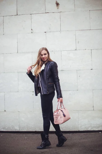 Porträt einer schönen blonden jungen kaukasischen Hipsterin in einer schwarzen Pelzjacke, die neben der weißen Backsteinwand mit rosa Tasche posiert. windiges Wetter Stadtporträt urban — Stockfoto