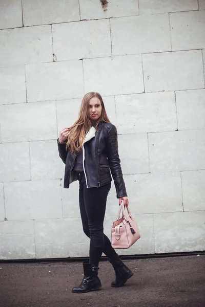 Ritratto di una bella bionda giovane donna caucasica hipster In una giacca di pelliccia nera in posa di moda accanto al muro di mattoni bianchi con borsa rosa. meteo città ritratto urbano — Foto Stock