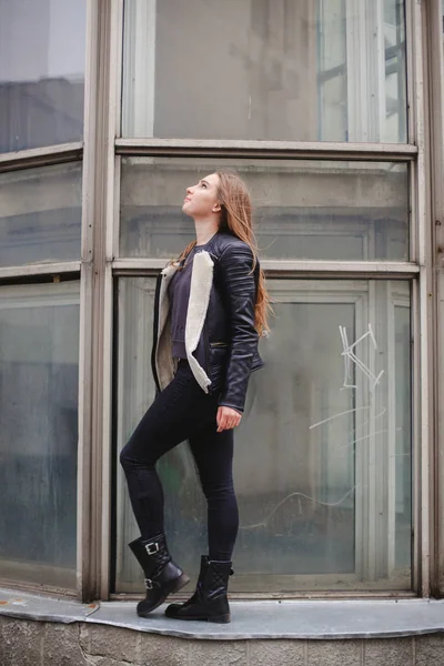 Porträtt av en vacker blond ung kaukasisk kvinna hipster i en svart pälsjacka poserar mode bredvid den blå glas byggnad. staden porträtt urban — Stockfoto