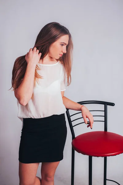 studio portrait of beautiful thin blonde caucasian young woman girl, isolated on white background posing in a white shirt and black office skirt. standing near red bar stool and doing fashion poses