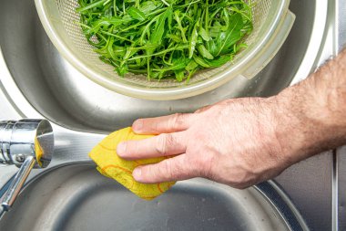 Cleaning the sink. The man wipes the silver sink with a cloth. The concept of care for cleanliness in the kitchen.