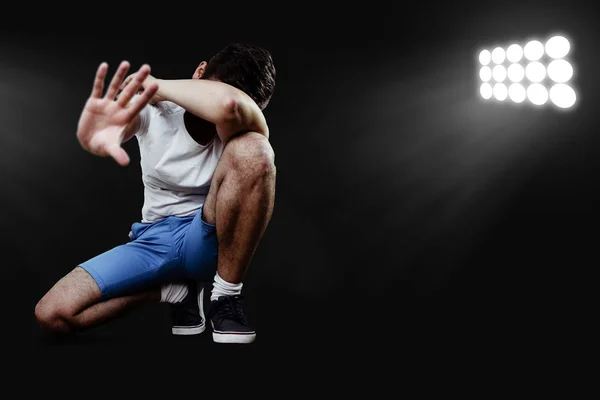 The huddled man covers his face with his hand and distracts people with his other hand. A sports man dressed in exhaustion squats on the ground in the light of a dark background.
