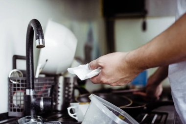 The man wipes clean clean dishes with a towel. Dirty dishes in the sink. No dishwasher. Concept of caring for dishes in the kitchen. The kitchen sink is full of dirty dishes.