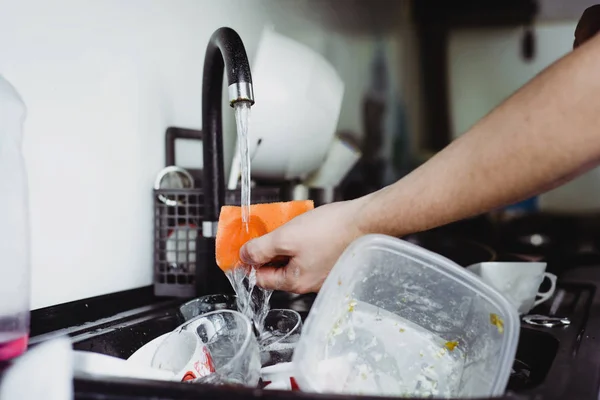 The man cleans dirty dishes with a dishcloth. Dirty dishes in the sink. No dishwasher. Concept of caring for dishes in the kitchen. The kitchen sink is full of dirty dishes.