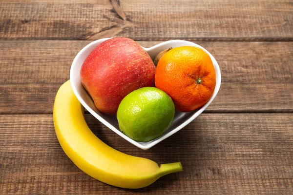 Heart-shaped bowl with fruit on wooden background. The concept of healthy eating, eating fruit. Bowl with apple, mandarin banana. A bowl filled with fruit. Fruit and vitamins for the heart.