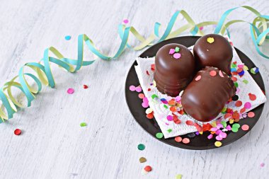 Çikolata kaplı küçük kremalı pastalar konfeti ve flamalarla bir yüzeyde dururlar. Çikolata marşmelovu parlak bir yüzeyde karnaval için dururlar.