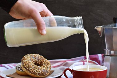 Sütü cam bir şişeden, arka planda koyu bir arka plan ve çöreklerle tahta bir fonda duran kahve dolu kırmızı bir fincana dökün.