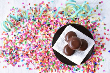 Çikolata kaplı küçük kremalı pastalar konfeti ve flamalarla bir yüzeyde dururlar. Çikolata marşmelovu parlak bir yüzeyde karnaval için dururlar.