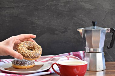Bir elinde donut, yanında koyu, ahşap arka planda süt ile karıştırılmış bir kahve.