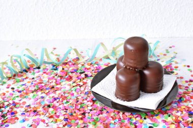 Çikolata kaplı küçük kremalı pastalar konfeti ve flamalarla bir yüzeyde dururlar. Çikolata marşmelovu parlak bir yüzeyde karnaval için dururlar.