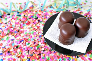 Çikolata kaplı küçük kremalı pastalar konfeti ve flamalarla bir yüzeyde dururlar. Çikolata marşmelovu parlak bir yüzeyde karnaval için dururlar.