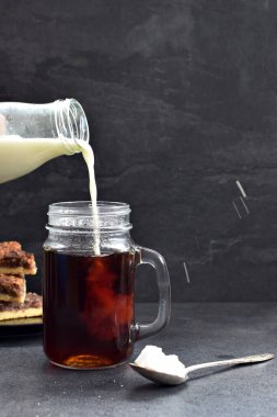 Fındık köşeli koyu bir arka fona karşı kahve dolu bir bardağa süt dökün - taze demlenmiş sıcak kahve içine süt akan koyu bir ayara karşı.