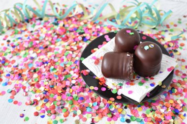 Çikolata kaplı küçük kremalı pastalar konfeti ve flamalarla bir yüzeyde dururlar. Çikolata marşmelovu parlak bir yüzeyde karnaval için dururlar.