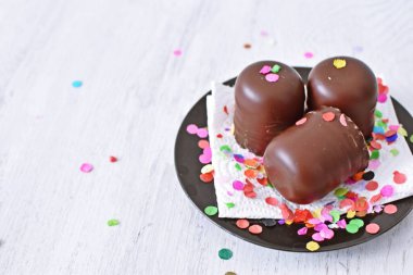 Çikolata kaplı küçük kremalı pastalar konfeti ve flamalarla bir yüzeyde dururlar. Çikolata marşmelovu parlak bir yüzeyde karnaval için dururlar.