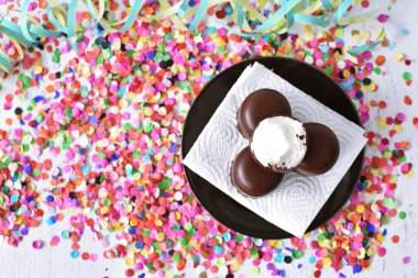 Çikolata kaplı küçük kremalı pastalar konfeti ve flamalarla bir yüzeyde dururlar. Çikolata marşmelovu parlak bir yüzeyde karnaval için dururlar.