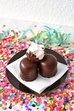 Çikolata kaplı küçük kremalı pastalar konfeti ve flamalarla bir yüzeyde dururlar. Çikolata marşmelovu parlak bir yüzeyde karnaval için dururlar.