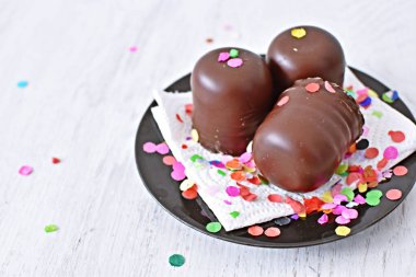 Çikolata kaplı küçük kremalı pastalar konfeti ve flamalarla bir yüzeyde dururlar. Çikolata marşmelovu parlak bir yüzeyde karnaval için dururlar.