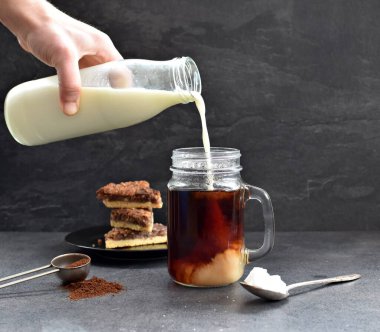 Fındık köşeli koyu bir arka fona karşı kahve dolu bir bardağa süt dökün - taze demlenmiş sıcak kahve içine süt akan koyu bir ayara karşı.