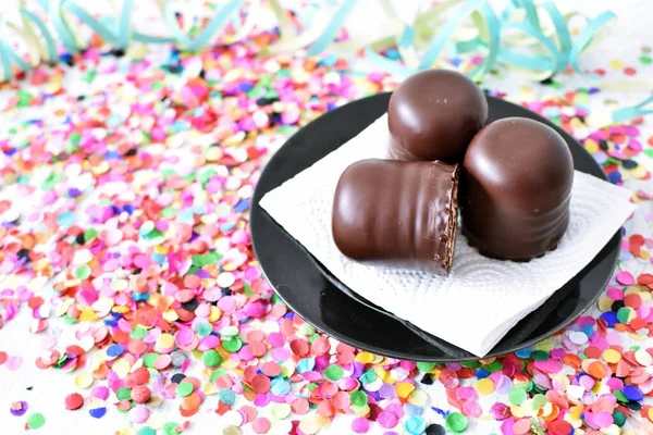 Çikolata kaplı küçük kremalı pastalar konfeti ve flamalarla bir yüzeyde dururlar. Çikolata marşmelovu parlak bir yüzeyde karnaval için dururlar.