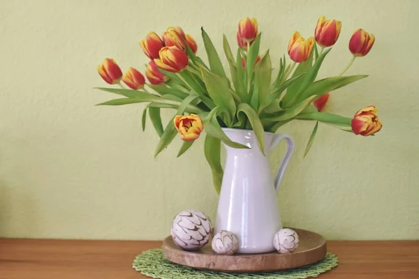 A white porcelain can as a vase stands in front of a green background with yellow-red tulips inside. Decoration for spring against a green background