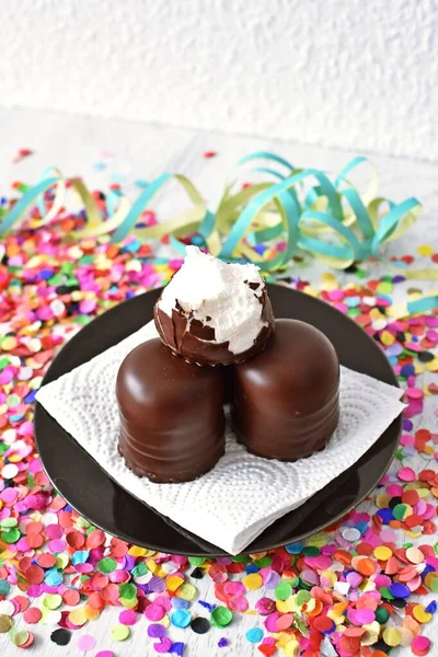 Çikolata kaplı küçük kremalı pastalar konfeti ve flamalarla bir yüzeyde dururlar. Çikolata marşmelovu parlak bir yüzeyde karnaval için dururlar.