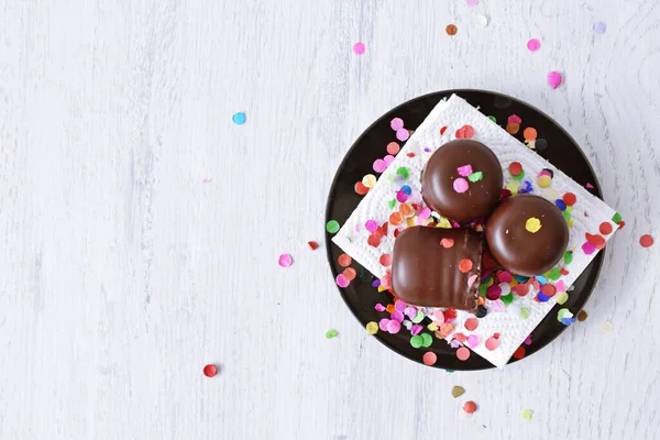 Çikolata kaplı küçük kremalı pastalar konfeti ve flamalarla bir yüzeyde dururlar. Çikolata marşmelovu parlak bir yüzeyde karnaval için dururlar.