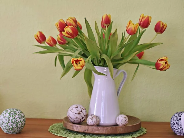 Vazo olarak beyaz bir porselen kutu içinde sarı-kırmızı laleler olan yeşil bir arka planın önünde duruyor. Yeşil arka planda bahar için dekorasyon