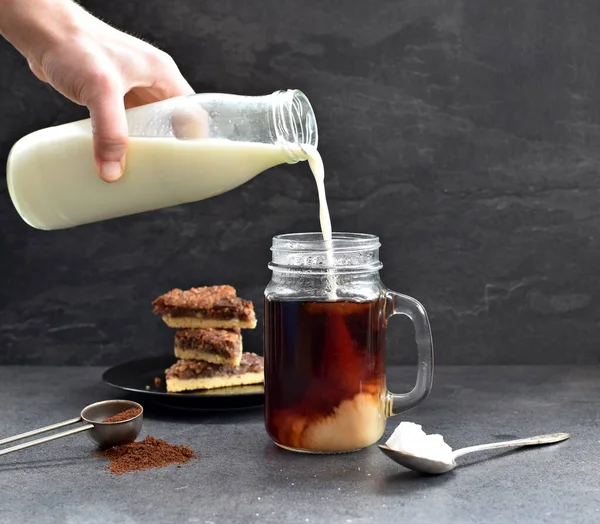 Fındık köşeli koyu bir arka fona karşı kahve dolu bir bardağa süt dökün - taze demlenmiş sıcak kahve içine süt akan koyu bir ayara karşı.