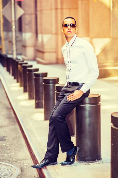 Young American Man wearing white shirt, black pants, leather shoes, sunglasses, sitting metal pillar on the street, relaxing, waiting for you. Filtered effec