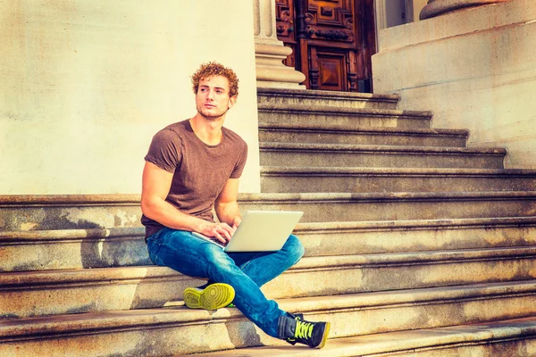 Молодой человек работает за ноутбуком, сидит на лестнице снаружи — стоковое фото