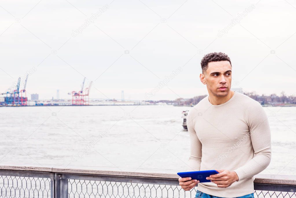 Young Handsome American Man traveling in New York, wearing light gray collarless, knit pullover sweater, standing by fence at harbor, holding blue tablet computer, reading, looking, thinking